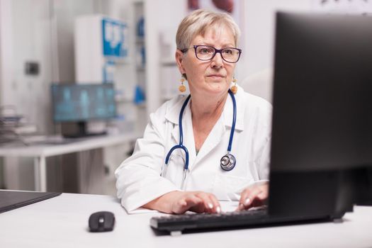 Mature doctor typing patient report on computer in hospital cabinet wearing stethoscope around the neck