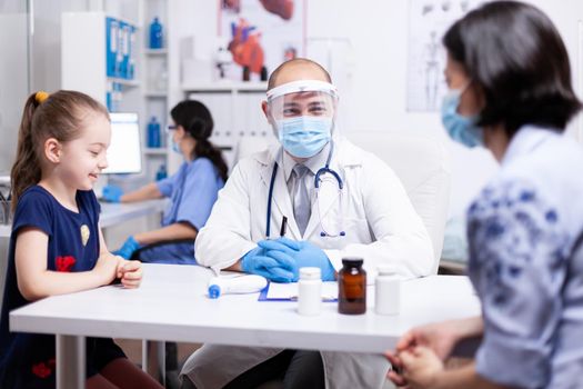 Pediatrician wearing face mask consulting kid during covid-19 in hospital office. Health doctor specialist providing health care services consultations treatment in protective equipment.