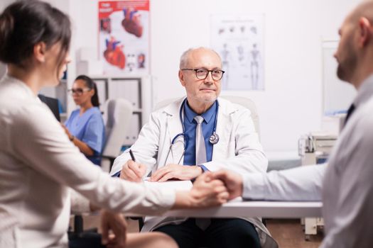 Senior doctor giving bad news to young couple during medical check in hospital office. Husband and wife holding hands. Nurse looking at patient x-ray.