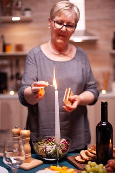 Candle burning on kitchen table for romantic dinner. Elderly woman waiting her husband for a romantic dinner. Mature wife preparing festive meal for anniversary celebration.