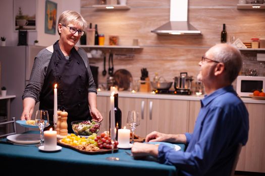 Senior couple celebration relationship having a romantic dinner in kitchen. Elderly old couple talking, sitting at the table in kitchen, enjoying the meal,