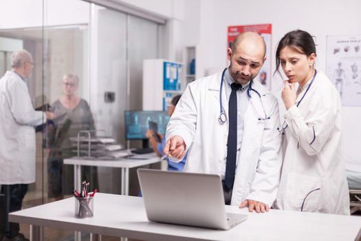 Team work of young doctors in hospital office wearing white coats and stethoscope looking at laptop. Senior medic with elderly aged patient on clinic corridor.