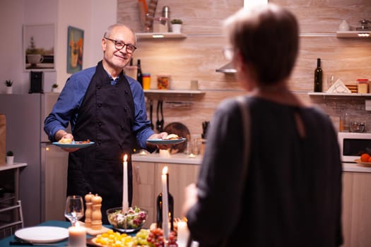 Senior man smiling at wife while holding plates with food food their romantic relationship anniversary. Elderly old couple talking, sitting at the table in kitchen, enjoying the meal,