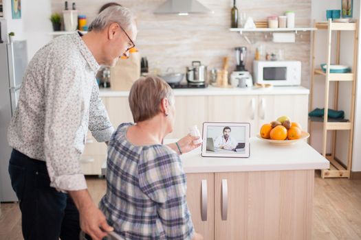 Senior couple listening doctor during telemedicine conference. Video conference with doctor using laptop in kitchen. Online health consultation for elderly people drugs ilness advice on symptoms, physician telemedicine webcam. Medical care internet chat