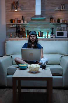 Woman freelancer in pijamas working late at night on laptop in front tv. Happy person in pijamas with eye mask sitting on sofa reading writing searching browsing on notebook using internet technology