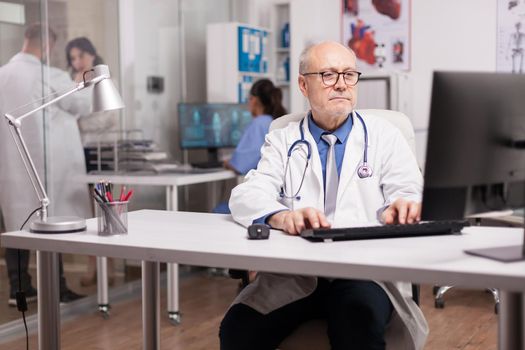 Senior doctor with stethoscope using pc in hospital cabinet while young medic wearing white coat checking report with patient on hospital corridor and nurse in blue uniform.