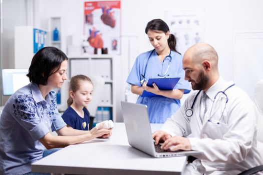 Doctor writing treatment on laptop during consultation of child and mother in hospital office. Healthcare physician specialist in medicine providing health care services treatment examination.