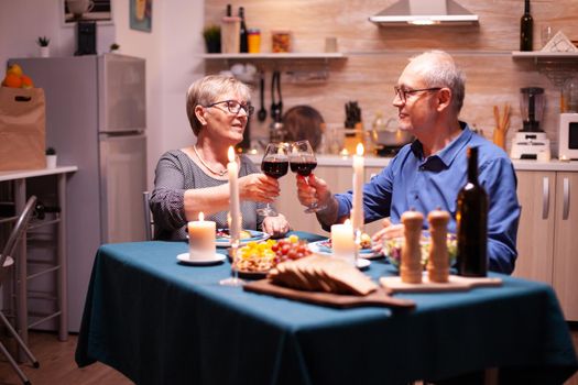 Cheerful old couple celebrating marriage with wine and festive dinner. Happy senior elderly couple dining together in the cozy kitchen, enjoying the meal, celebrating their anniversary.