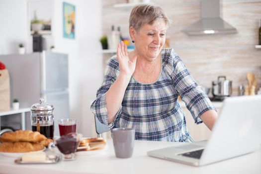 Senior woman drinking coffee and waving during video conference with family in kitchen while having breakfast. Elderly person using internet online chat technology video webcam making a video call connection camera communication conference call