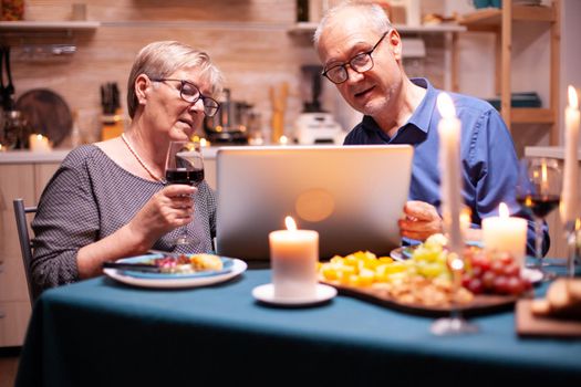 Aged man and woman shopping online using laptop in kitchen during festive dinner. Woman holding glass of wine. Elderly people sitting at the table browsing, searching, using laptop, technology, internet, celebrating their anniversary in the dining room.