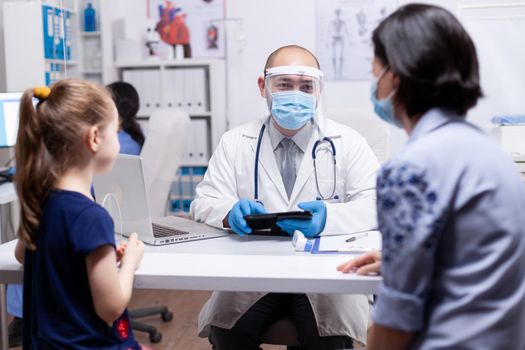 Doctor talking about child treatment with mother during coronavirus pandemic wearing face mask and holding tablet pc. Pediatrician specialist with protection mask providing health care.