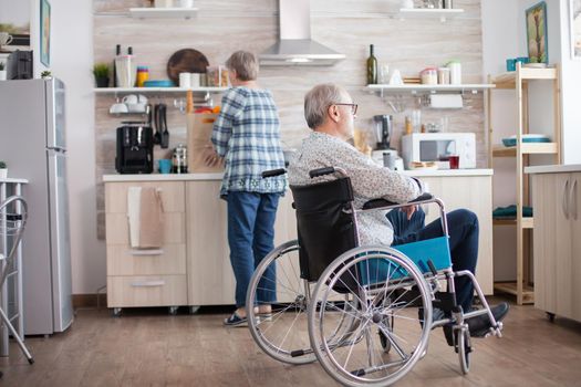 Pensive disabled elderly person in wheelchair looking on the window from the kitchen. Disabled man sitting in wheelchair in kitchen looking through window while wife is preparing breakfast. Invalid, pensioner, handicapped, paralysis.