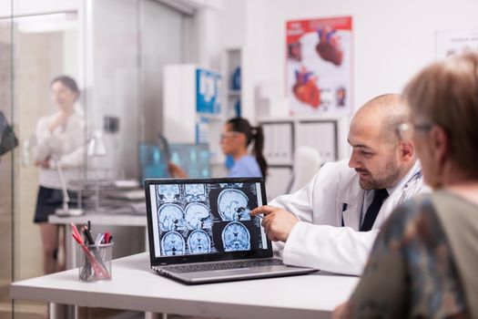 Brain surgeon pointing at ct scan on laptop in hospital office discussing with elderly patient about intervention. Nurse holding x-ray image in the background. Young sick woman in clinic corridor.