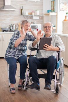 Cheerful senior woman waving on video conference in the kitchen. Disabled senior man in wheelchair and his wife having a video conference on tablet pc in kitchen. Paralyzed old man and his wife having a online conference.