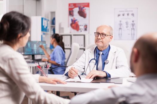 Senior doctor with grey hair during patient examination in hospital office. Disabled young man in wheelchair with wife in clinic cabinet for medical check.