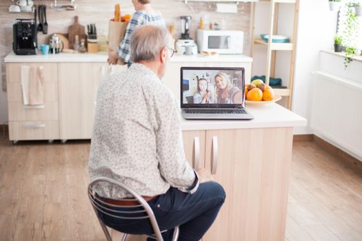 Senior man talking with niece on video call in the kitchen. Happy senior man during a video conference with family using laptop in kitchen. Online call with daughter and niece. Elderly person using modern communication online internet web techonolgy.