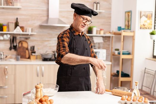 Experienced senior chef cooking healthy balanced food in home kitchen. Retired senior chef with bonete and apron, in kitchen uniform sprinkling sieving sifting ingredients by hand.