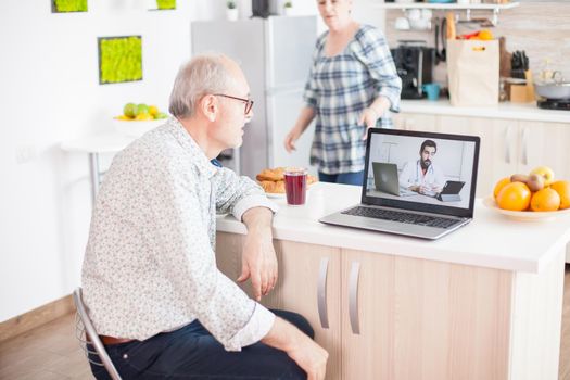 Senior retired man in video conference with doctor using laptop in kitchen. Online health consultation for elderly people drugs ilness advice on symptoms, physician telemedicine webcam. Medical care internet chat