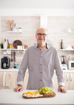 Happy elderly aged man smiling looking at camera in kitchen with tasty cheese appetizier and delicious grapes on table top.