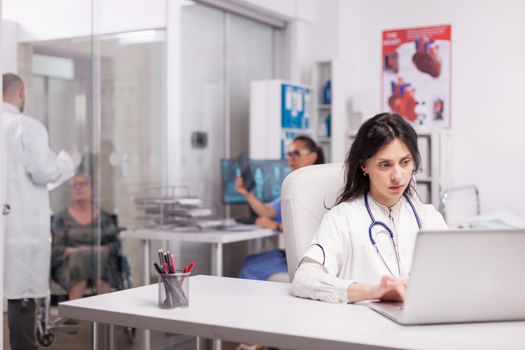 Young woman therapist in medical centre using laptop, disabled senior woman in wheelchair discussing with doctor on clinic corridor and nurse in blue uniform holds x-ray image.