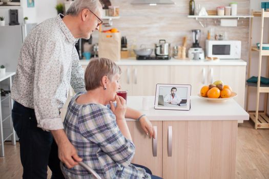 Worried senior woman in medical check with her husband next. Video conference with doctor using laptop in kitchen. Online health consultation for elderly people drugs ilness advice on symptoms, physician telemedicine webcam. Medical care internet chat