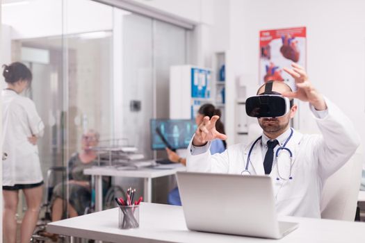 Young physician using virtual reality goggles in hospital office. Invalid senior woman in wheelchair talking with female doctor on clinic corridor. Nurse in blue uniform holding patient x-ray.