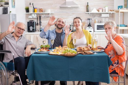 Disabled senior man in wheelchair and his family waving at camera during lunch in kitchen. Mature parents. Happy daughter and son.