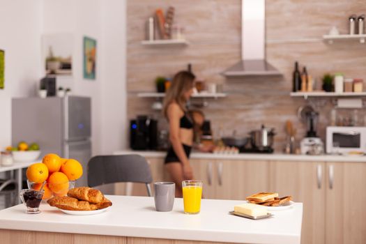Glass with fresh orange juice in kitchen during breakfast with sexy housewife in the background. Young sexy seductive blode lady with tattoos drinking healthy, natural juice.