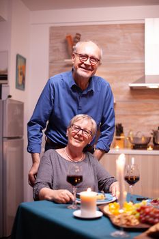 Disabled senior woman in wheelchair and husband looking at camera. Happy cheerful senior elderly couple dining together in the cozy kitchen, enjoying the meal, celebrating their anniversary.