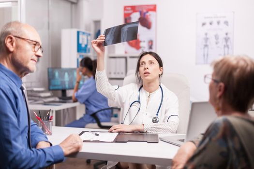 Female doctor looking at senior patient x-ray during consultation after surgery. Medic examining radiography in hospital office. Nurse working on computer.