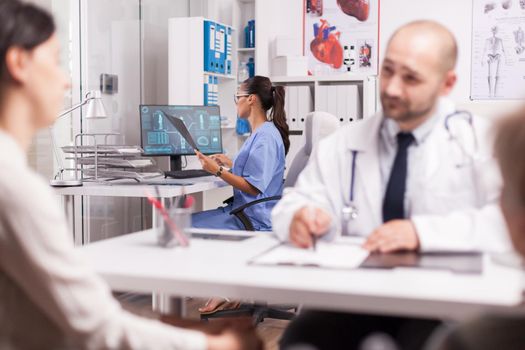 Nurse is examining patient x-ray in hospital office. Doctor with white coat and stethoscope explaining treatment to young woman and her disabled mother in wheelchair.