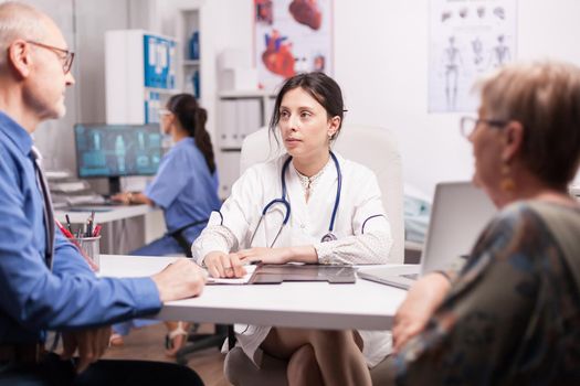 Young woman doctor in hospital office during consultation of old married couple wearing white coat and stethoscope. Nurse in blue uniform working on computer.