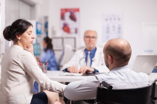 Disabled young man in wheelchair and his wife during consultation with senior doctor in hospital office. Elderly aged doctor wearing white coat and stethoscope. Nurse with blue uniform in the background.