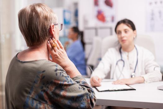 Mature woman suffering neck pain during consultation with doctor in hospital cabinet. Senior patient examination.