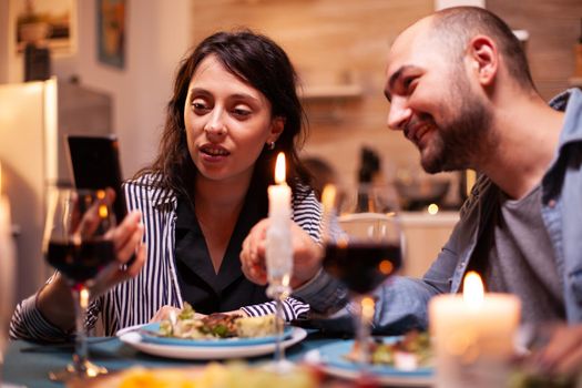 Couple surfing on internet using smartphone during romantic dinner in kitchen. Adults sitting at the table in the kitchen browsing, searching, using smartphones, internet, celebrating anniversary.