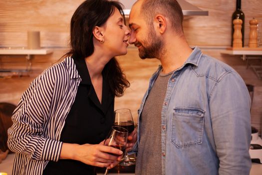 Woman giving affection to husband during romantic date and holding glass of wine. Adult couple at home, drinking red wine, talking, smiling, enjoying the meal in dining room.