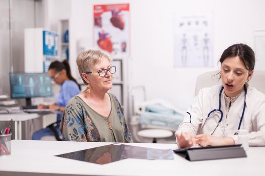 Doctor using tablet computer while examining senior woman in hospital office. Medic discussing diagnosis with patient about her illness.