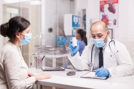 Doctor with face mask against coronavirus outbreak holding pills bottle while writing prescription for sick woman. Nurse wearing blue uniform working on computer.