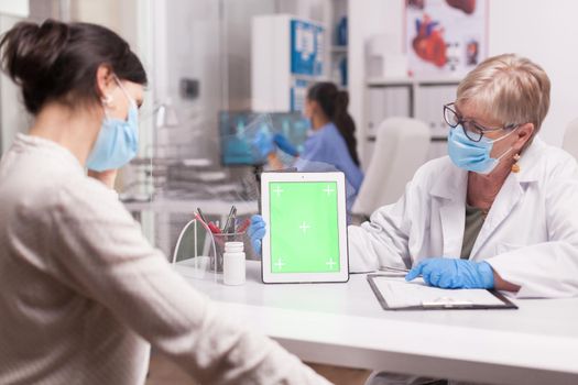 Doctor with face mask against coronavirus holding tablet computer with green screen during consultation with sick patient.