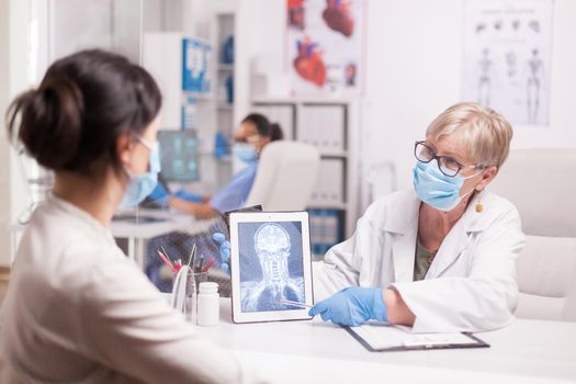 Senior doctor with mask against covid-19 holding tablet computer with brain x-ray showing patient head trauma. Medic discussing treatment with sick woman.