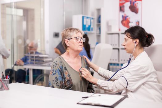 Doctor checking old woman heart beat with stethoscope in hospital office. Disabled senior man in wheelchair talking with medic in clinic corridor.