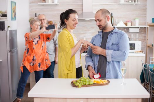 Young man proposing wife in fron of her parents during family lunch. Happy woman holding glass of wine.