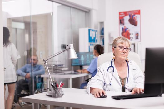 Elderly aged woman doctor reading expertise report on computer while young therapist looking at handicapped patient in wheelchair and nurse wearing blue uniform.