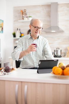 Elderly man surfing on internet using tablet computer during breakfast in kitchen. Senior person with tablet portable pad PC in retirement age using mobile apps, modern internet online information technology with touchscreen