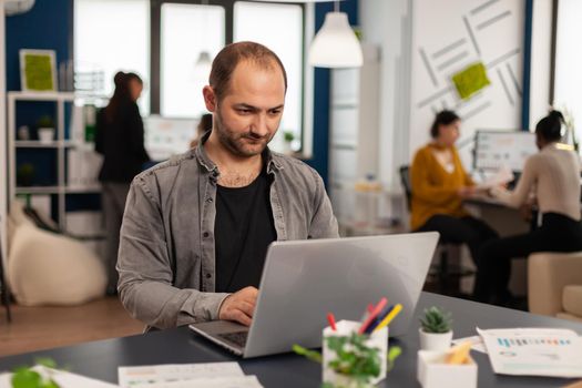 Man authentic manager sitting at table in modern startup business office sending mails typing on laptop. Business employee working in financial company with diverse team