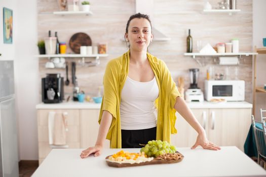 Cheerful wooman looking at camera in home kitchen with delicious variety of cheese and grapes on wooden plate