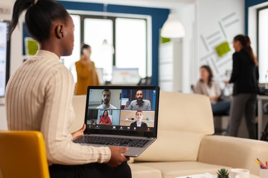 Tired black woman talking with colleague using video call holding smartphone with wiraless headset sitting on couch in business office. Diverse team planning new financial project in modern company