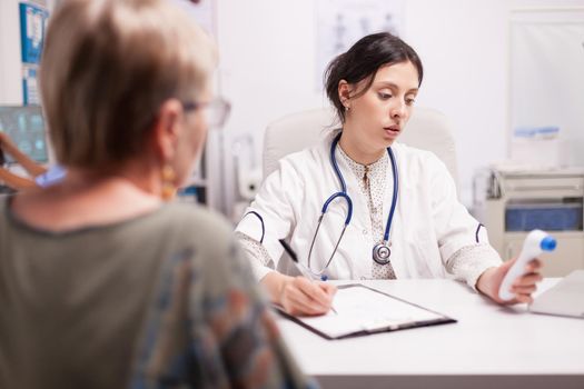 Medic writing senior patient diagnosis after measuring body temperature with thermometer gun in hospital office. Doctor with stethoscope wearing white coat.