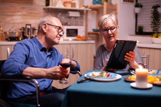 Elderly woman browsing on tablet pc and disabled husband in wheelchair holding wine glass. Imobilized handicapped senior husband browsing on phone enjoying the festive male, drinking a glass of red wine.