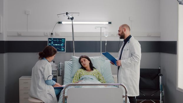 Specialist doctors monitoring sick woman discussing sickness treatment while working in hospital ward. Patient resting in bed explaining symptom disease during recovery appointment